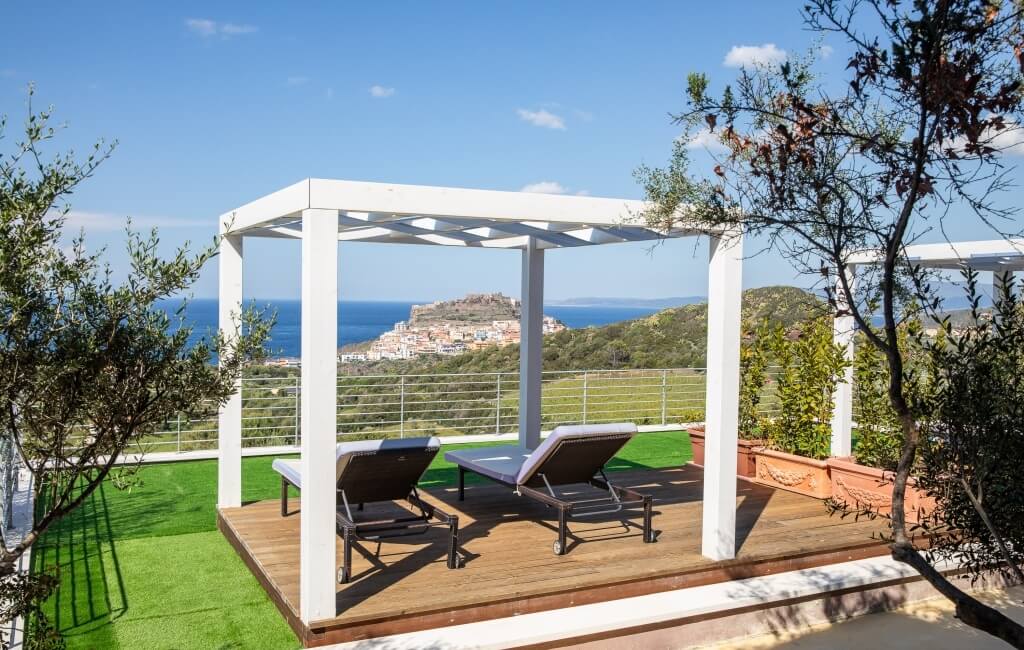 Weißer Pergola mit Liegestühlen auf Holzdeck, Blick auf den Ozean und eine charmante Stadt auf einem Hügel.