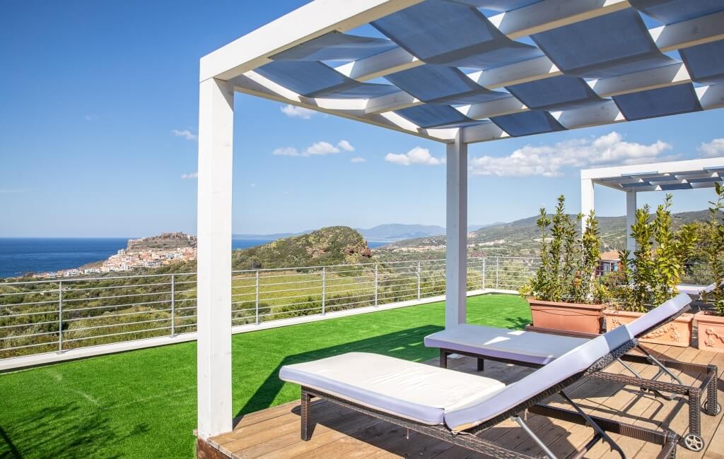 Ein weißer Pavillon mit zwei Liegestühlen auf einer Holzterrasse mit Blick auf das Meer und die Berge im Hintergrund.