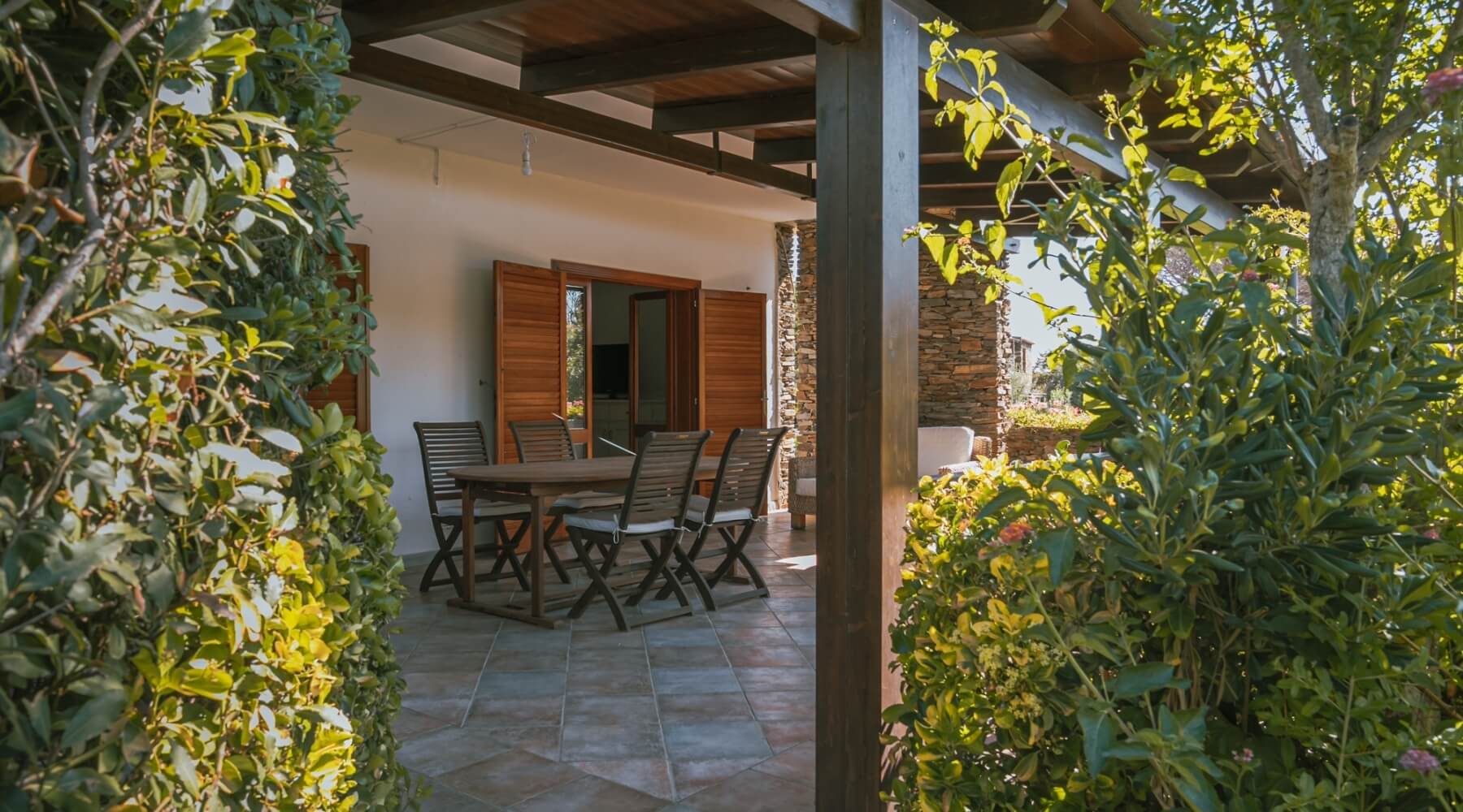 Patio con tavolo e sedie in legno, circondato da vegetazione.