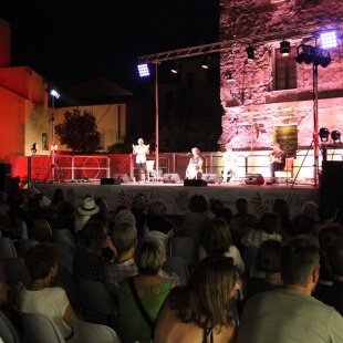 Palco all'aperto con artisti, pubblico seduto e illuminazione.