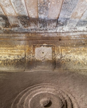 Interno con pareti ruvide, soffitto decorato e apertura centrale.