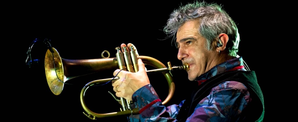 Uomo con capelli grigi suona uno strumento a fiato, sfondo nero.