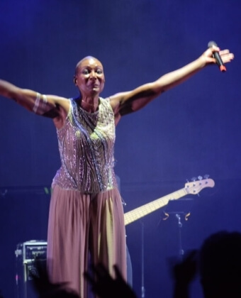 Donna calva sul palco con microfono, indossa top di paillettes.