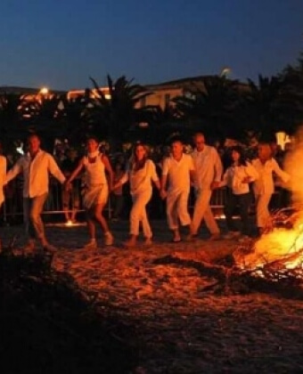 Gruppo di persone in bianco danza attorno a un falò sulla sabbia.