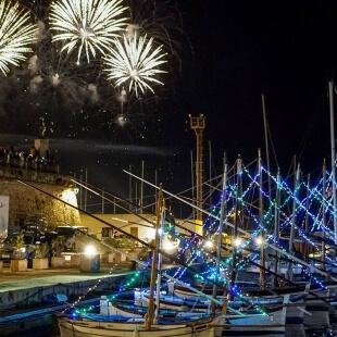Porto notturno con barche illuminate e fuochi d'artificio sopra.