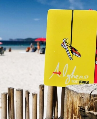 Cartello giallo con scarpe appese, spiaggia e persone sullo sfondo.