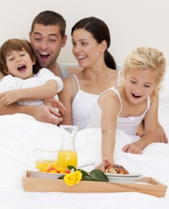 Una famiglia a letto con un vassoio di colazione. Una ragazza prende un pasticcino.