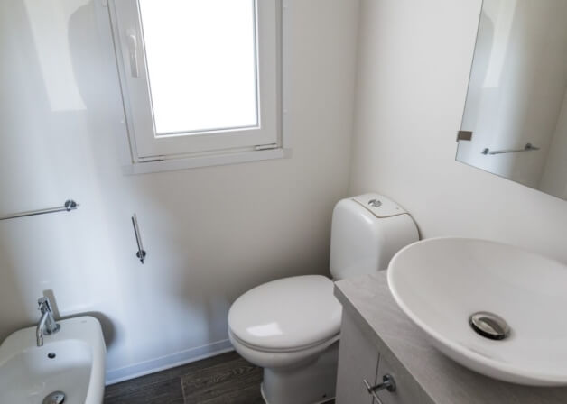 Bagno con WC, bidet, lavabo, finestra e pavimento in piastrelle effetto legno.