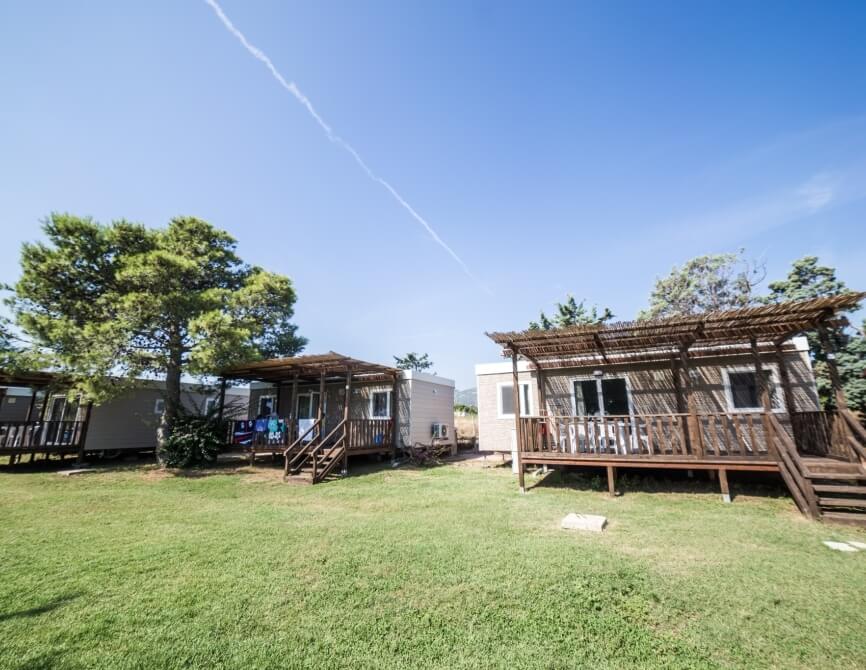 Case mobili con verande su un'area verde, alberi sullo sfondo.