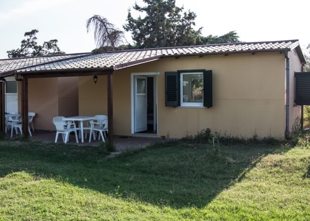 Bungalow beige con patio coperto, sedie bianche e alberi circostanti.