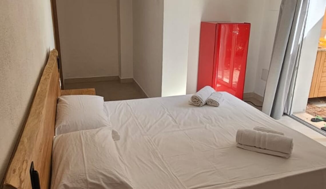 Letto con biancheria bianca e frigorifero rosso accanto.