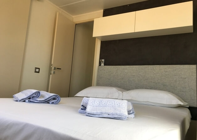 Letto con lenzuola bianche, testiera grigia e asciugamani a righe.