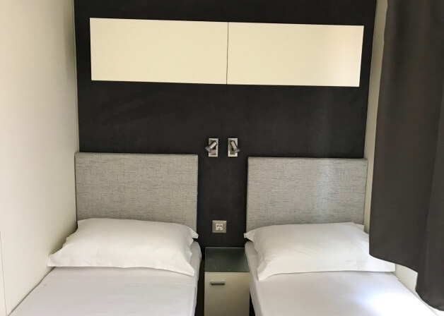 Camera d'hotel con due letti, testate grigie e armadietti bianchi.