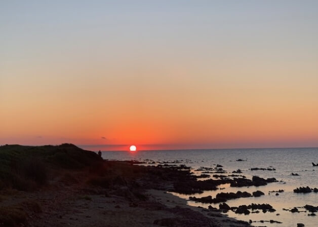 Tramonto arancione su acqua, figura sulla riva con rocce scure.