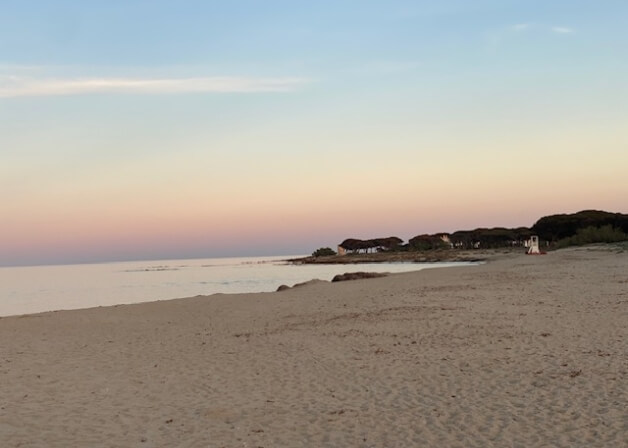 Spiaggia sabbiosa con acqua calma, albe rosa e arancioni, e torretta di salvataggio.