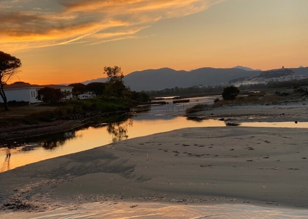 Paesaggio naturale al tramonto con acqua, colline e insediamento.