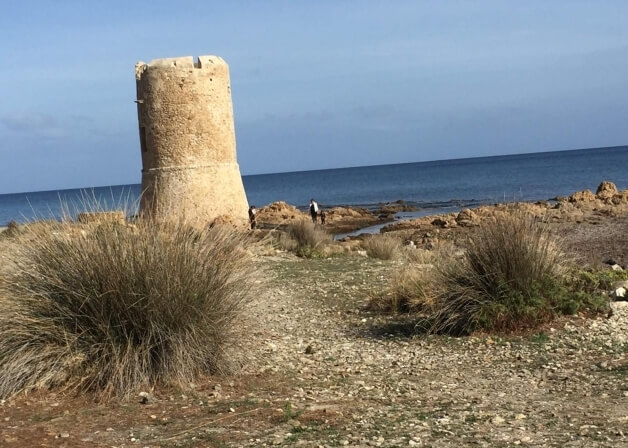 Torre di pietra su sfondo roccioso, mare e cielo blu.