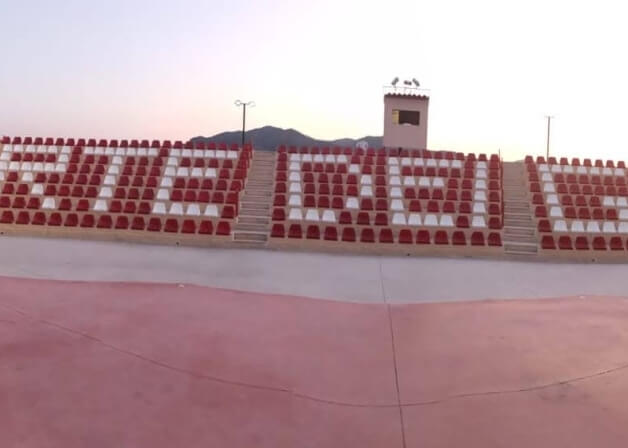 Stadio con sedie rosse e bianche che formano "FIEL DEL FORTIN", torre con bandiere e montagne sullo sfondo in una giornata serena.