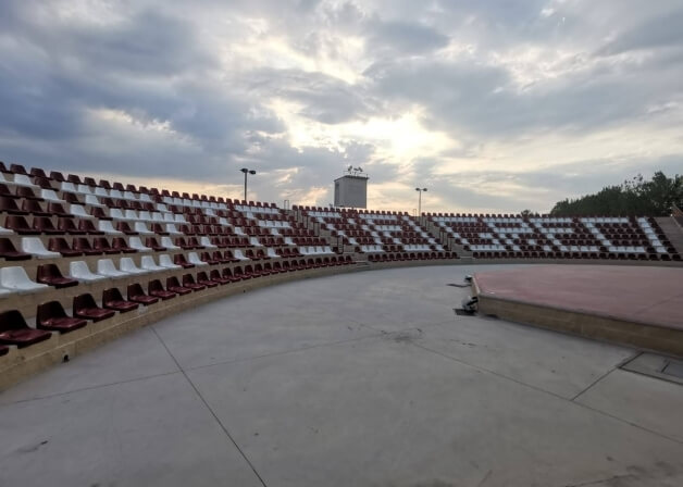 Un grande anfiteatro all'aperto con posti a sedere rossi e bianchi, un'area centrale e un cielo nuvoloso.