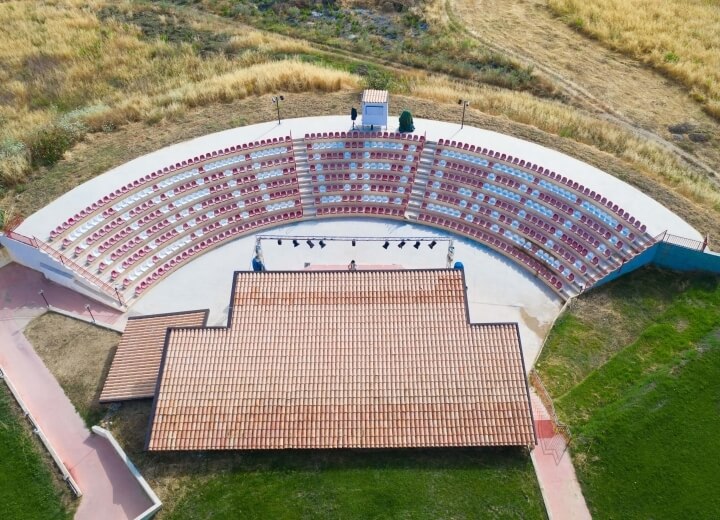 Vista aerea di un anfiteatro all'aperto con sedili rossi e bianchi in un paesaggio semi-arido con alberi sparsi.