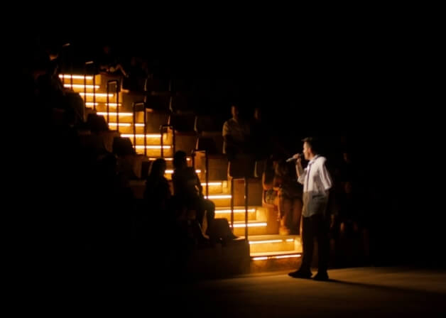 Un uomo canta in un microfono su un palco illuminato, con pubblico seduto in alto in un'atmosfera emozionante.