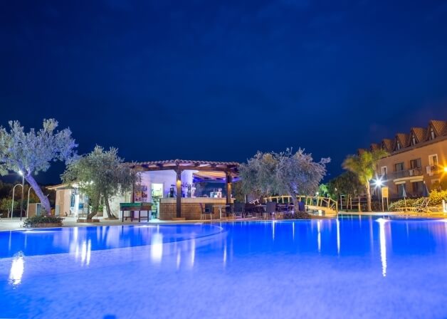 Una piscina serena di notte, riflettendo luci, con alberi e un bar all'aperto in un'atmosfera rilassata e tranquilla.