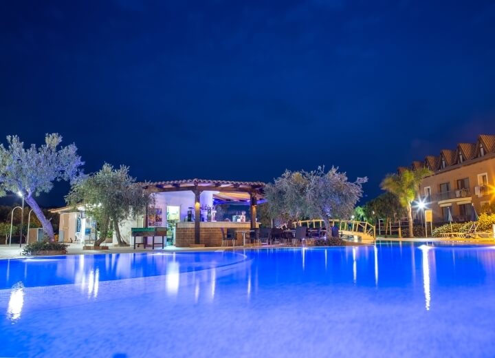 Scena serena di una piscina illuminata di blu di notte, con un bar e un edificio sullo sfondo in un resort.