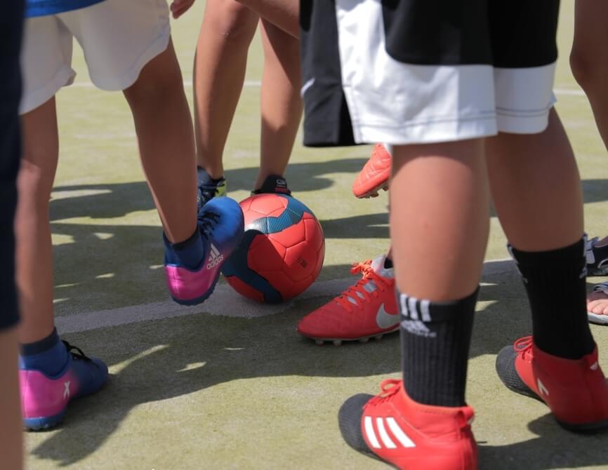 Un gruppo di gambe e piedi di bambini con un pallone da calcio rosso su un campo verde, indossando scarpe e calze colorate.