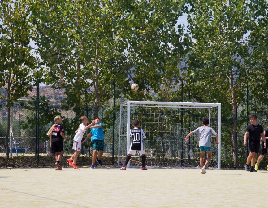 Un gruppo di bambini gioca a calcio all'aperto, con un goal bianco e alberi sullo sfondo, trasmettendo gioia e amicizia.