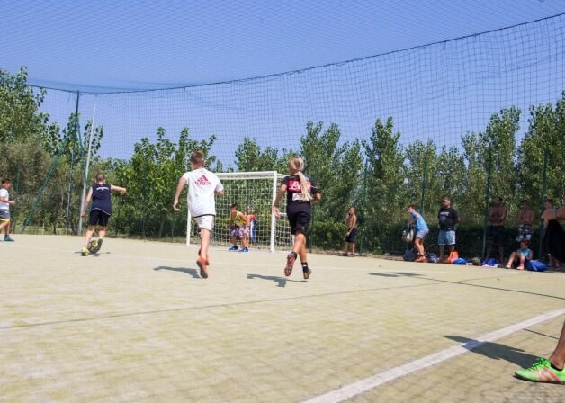 Una vivace scena di calcio con bambini che giocano su un campo soleggiato, circondati da alberi verdi e un cielo blu.