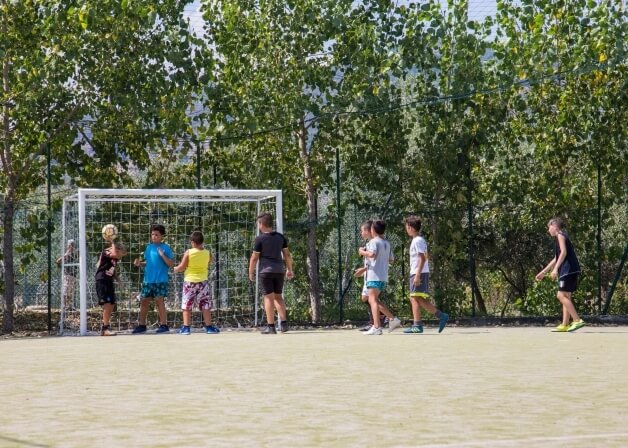 Un gruppo di otto bambini gioca a calcio su un campo all'aperto, vicino a un goal, alberi e una recinzione.