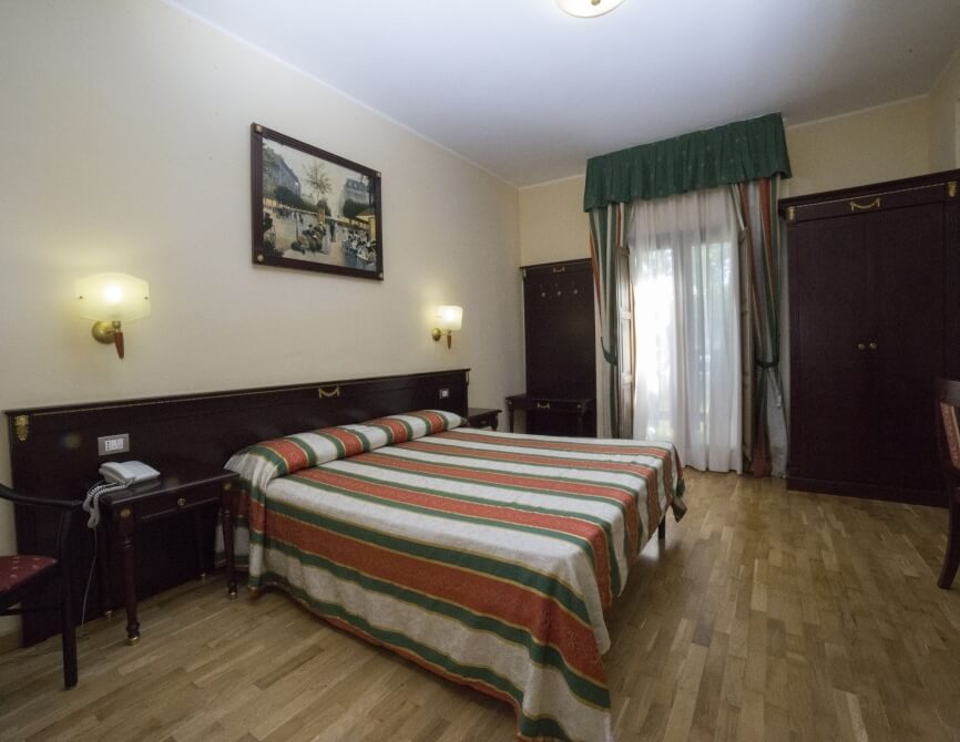 Camera d'albergo con letto grande, comforter a righe rosse e verdi, scrivania, armadio scuro e luce naturale dalla finestra.