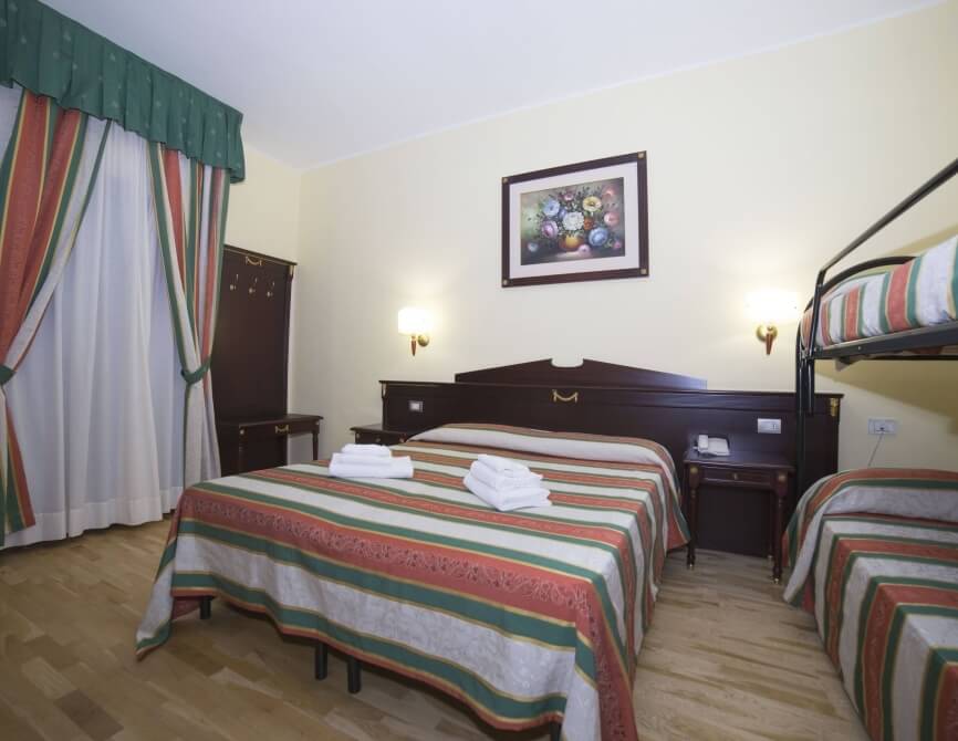 Camera d'hotel con due letti, testiere in legno scuro, biancheria bianca e comforter a righe, con tende bianche e pavimento in leg