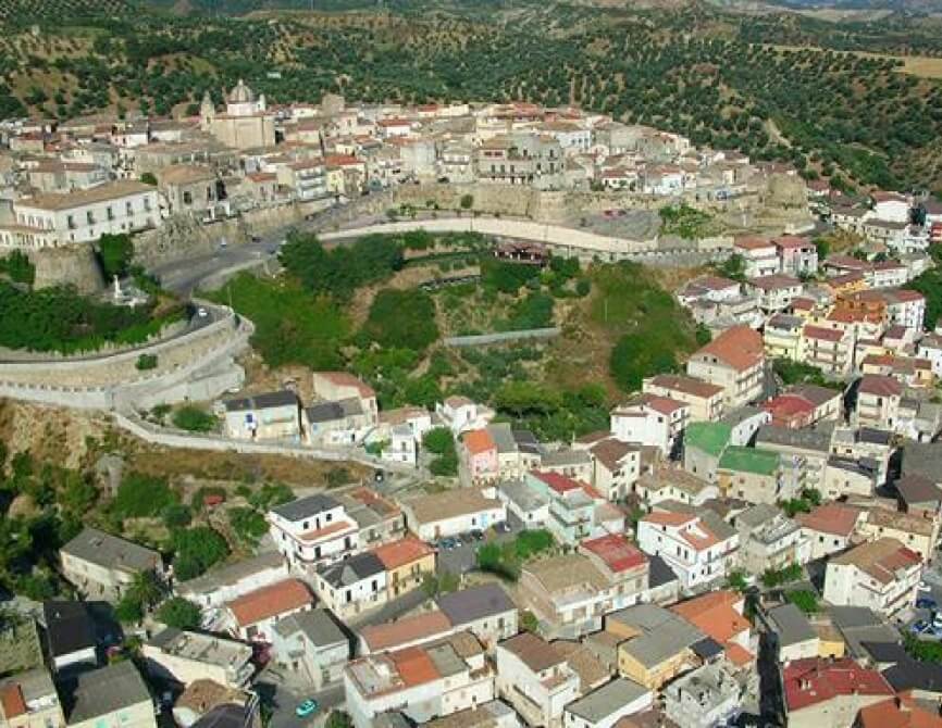 Vista aerea di un piccolo paese in una valle, con edifici bianchi e tetti rossi, circondato da colline verdi.