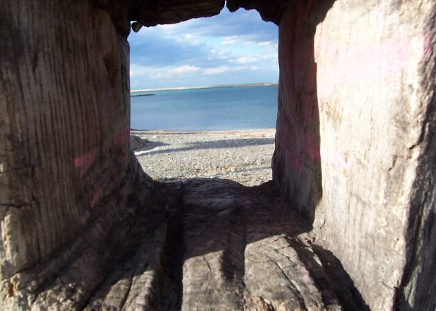 Un'apertura rettangolare in un muro di pietra incornicia una vista serena di un mare e una spiaggia rocciosa.