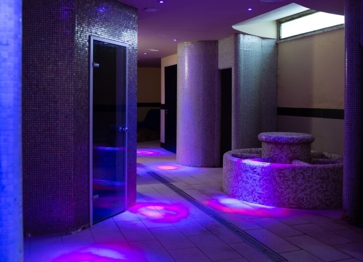 Interno di una spa lussuosa con colonne, piastrelle mosaico e luci viola e rosa che creano un'atmosfera rilassante.