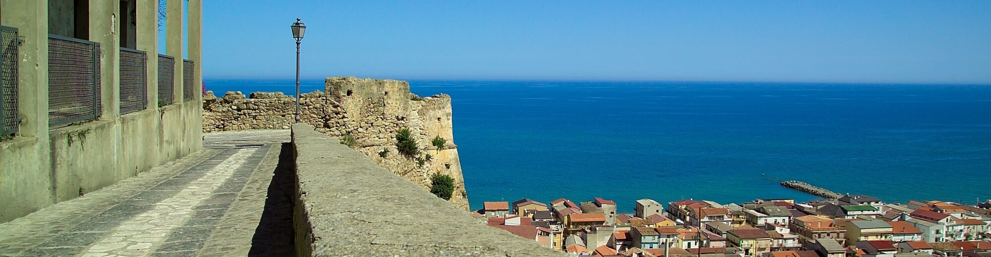 Sentiero in pietra con muro, lampione, città pittoresca e mare blu sotto un cielo sereno.