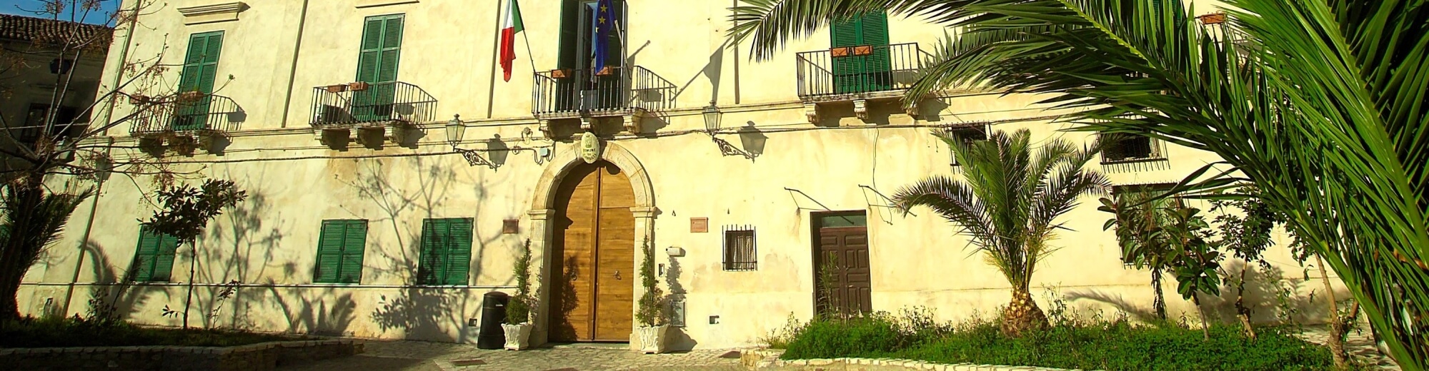 Edificio a due piani giallo chiaro con persiane verdi, balcone, bandiere francese e italiana, piante e alberi.