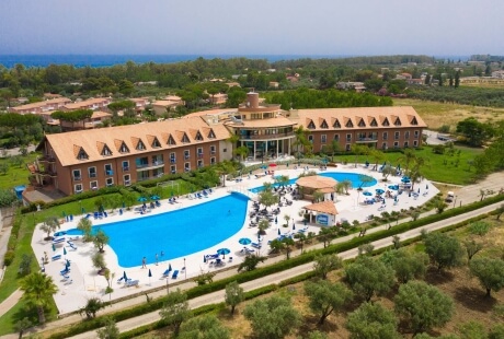 Vista aerea di un resort con piscine, palme e un edificio centrale con 14 finestre a dormer in un ambiente soleggiato.