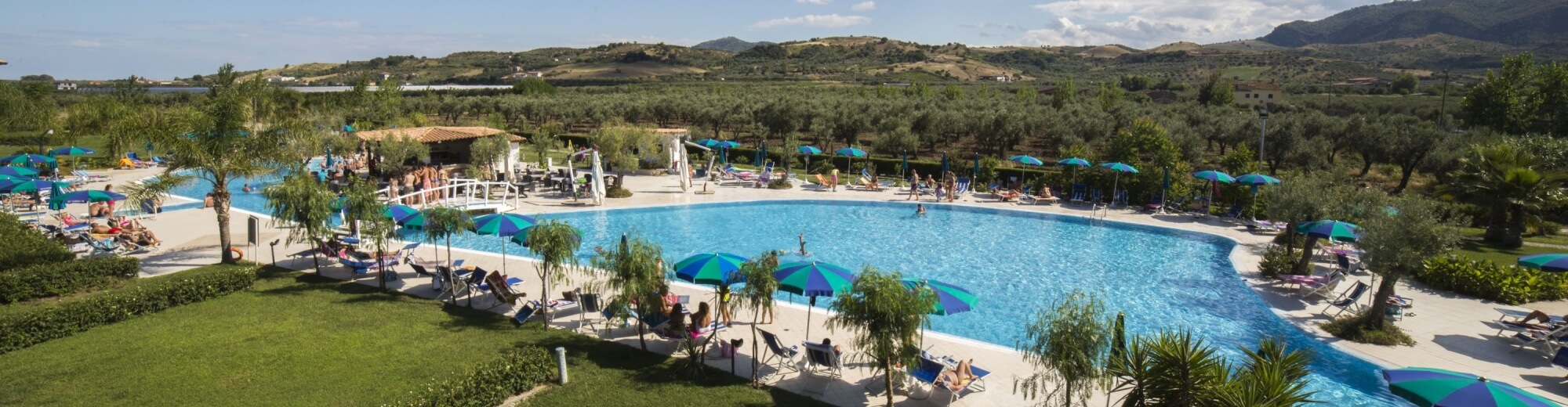 Una scena serena di un'area piscina con acqua blu, sdraio, ombrelloni e colline verdi sullo sfondo.