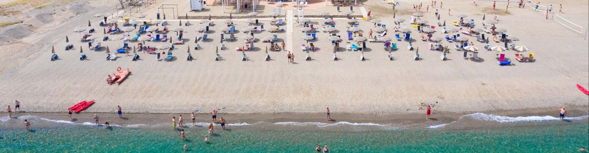 Una scena di spiaggia affollata con persone in acqua, sdraio, ombrelloni e kayak sotto il sole.