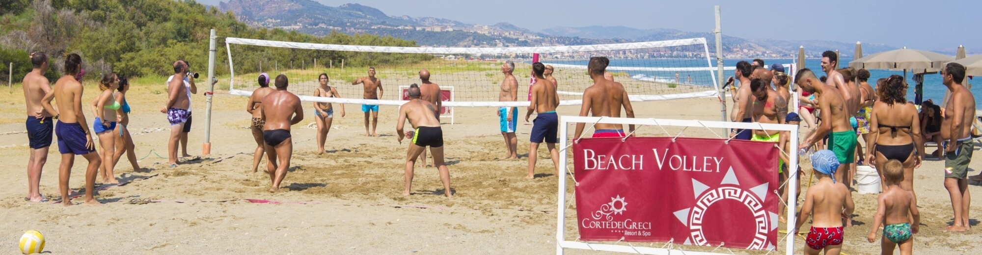 Una vivace scena di spiaggia con persone che giocano a pallavolo, circondate da sabbia, mare e montagne sotto un cielo blu.