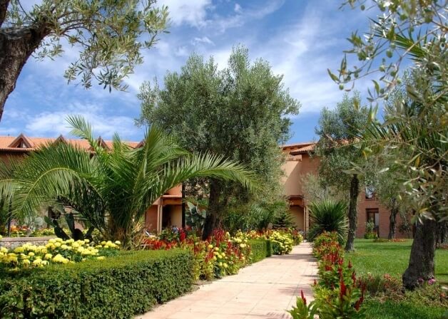 Un sentiero di giardino sereno conduce a un grande edificio color sabbia, circondato da piante e fiori vivaci sotto un cielo blu.