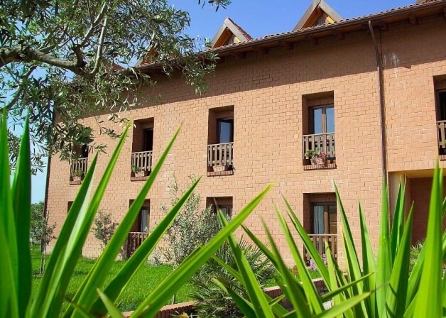 Edificio in mattoni beige a tre piani con finestre e balconi, circondato da erba verde e cielo blu.