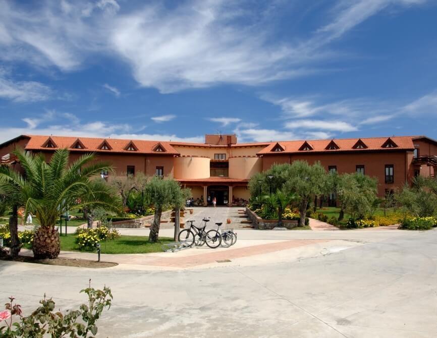 Edificio con tetto rosso, circondato da verde, biciclette parcheggiate e cielo blu con nuvole bianche. Atmosfera serena.