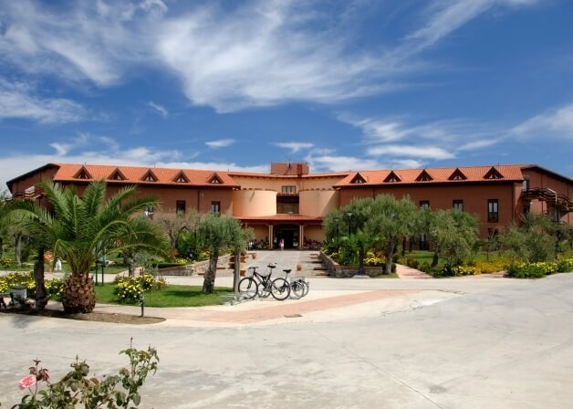 Edificio a due piani con tetto in tegole rosse, cortile, giardino curato e biciclette parcheggiate, sotto un cielo blu.