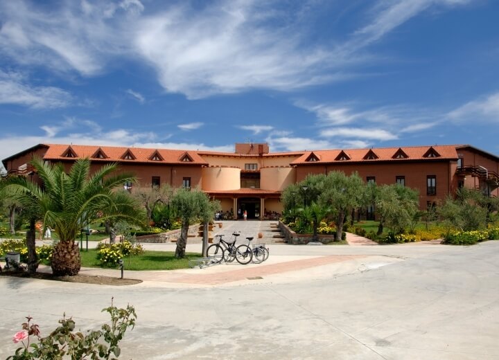 Edificio tan con tetto rosso, circondato da verde, biciclette parcheggiate, cielo blu sereno, atmosfera di relax.