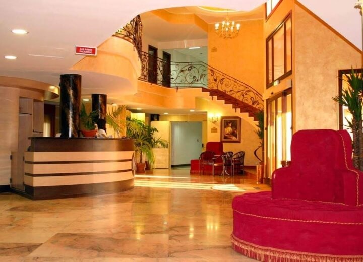 Lobby di hotel accogliente con pareti gialle, pavimento marmoreo, poltrona rossa, reception e piante decorative.