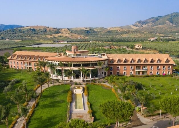 Un grande hotel con architettura moderna e rustica, circondato da un paesaggio sereno con fontana e colline.