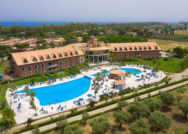 Complesso di resort con piscine, edifici a tetto rosso e persone che si rilassano, circondato da alberi e cielo nuvoloso.